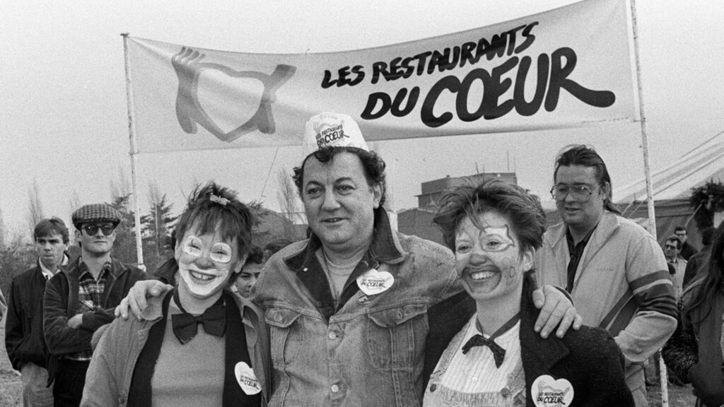 photographie de Coluche avec des bénévoles des Restos du Cœur